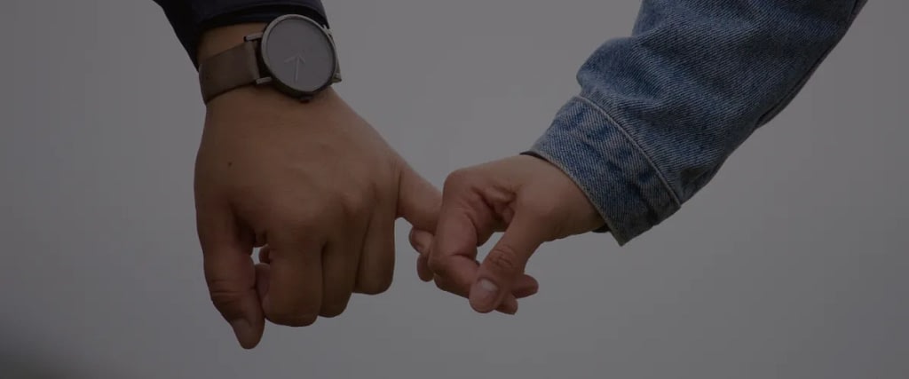 Two hands touch pinkies in a pinky-promise gesture against a neutral background, one wearing a watch.