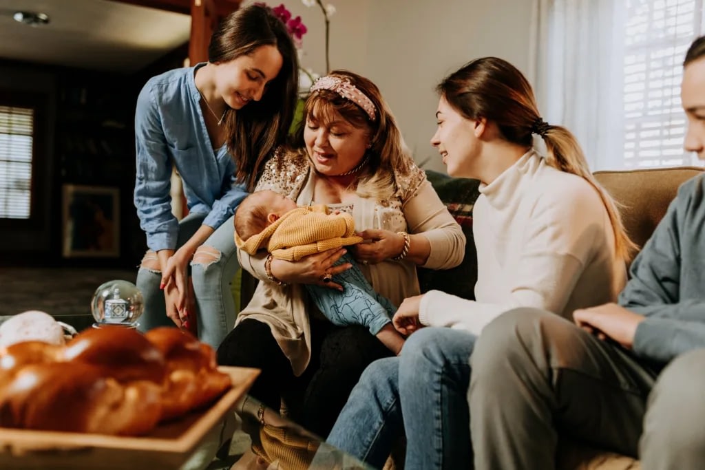 Women sit on a couch smiling at a newborn held by the central woman; pastries on a table in the foreground.