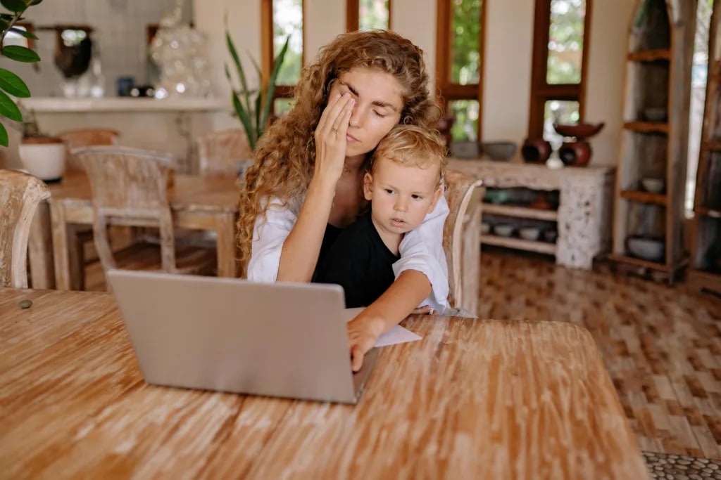 Mom and young child sit at a wooden dining table with a laptop, in a bright kitchen setting; the mother rests her hand on her face.",
