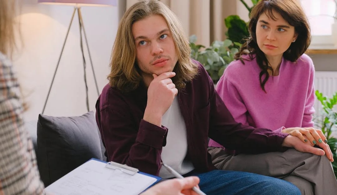 Couple in a therapy session sitting on a sofa; the man rests his chin thoughtfully while the woman looks on.