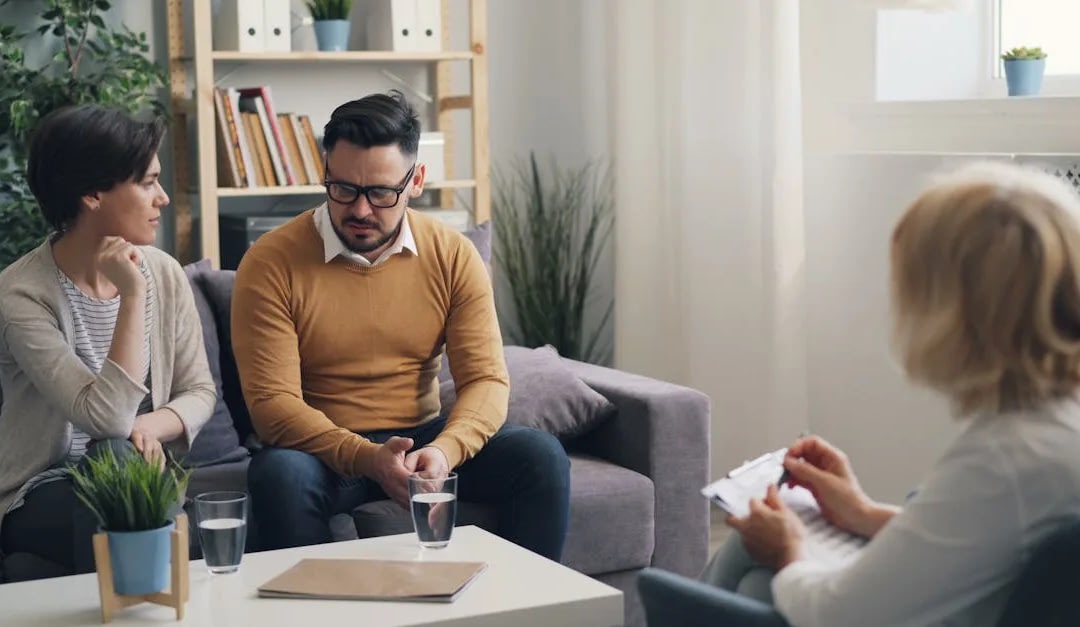 A man and a woman sit on a couch, talking with a clinician who holds a clipboard in a bright living-room therapy session.