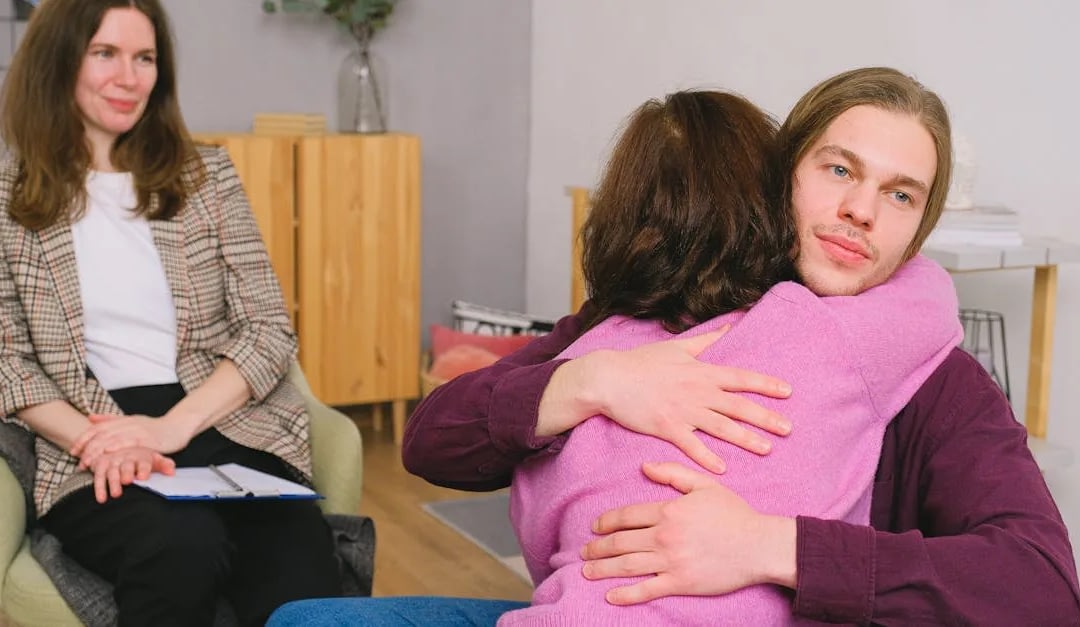 Therapist observes a couple hugging in a counseling room, clipboard in hand.