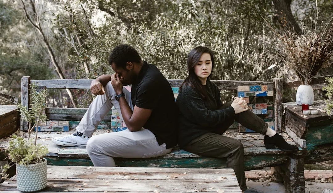 Two people sit back-to-back on a weathered wooden bench in a garden, both looking somber and lost in thought.