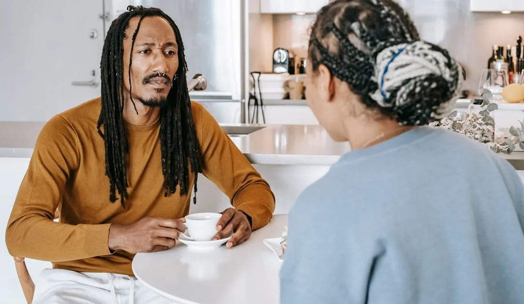 Man with dreadlocks in a brown shirt sits at a white table, holding a cup and talking to a woman with braided hair seen from behind in a bright kitchen.