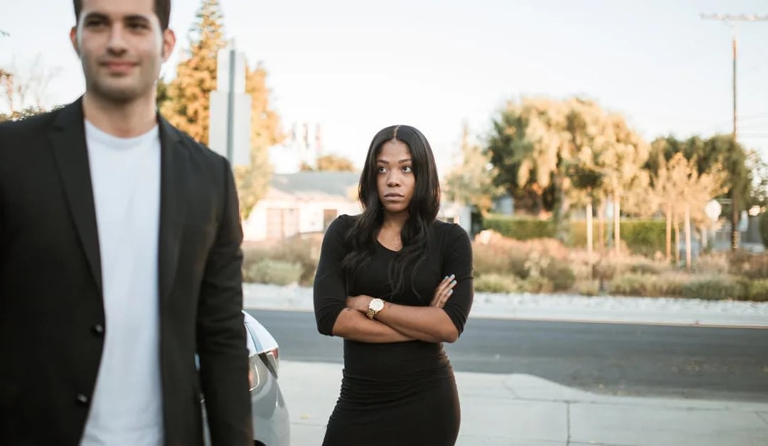 Woman with arms crossed on a sidewalk beside a parked car; man in a blazer is visible in the foreground, out of focus. Assumptions are being visually implied