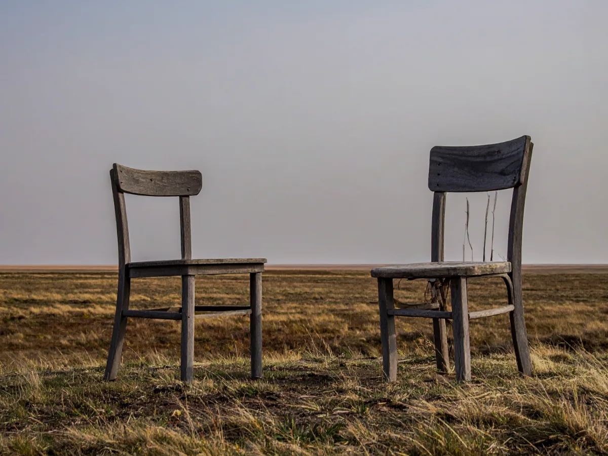 5 Two weathered wooden chairs sit in a dry field under a pale blue sky, one intact and the other with a broken back.