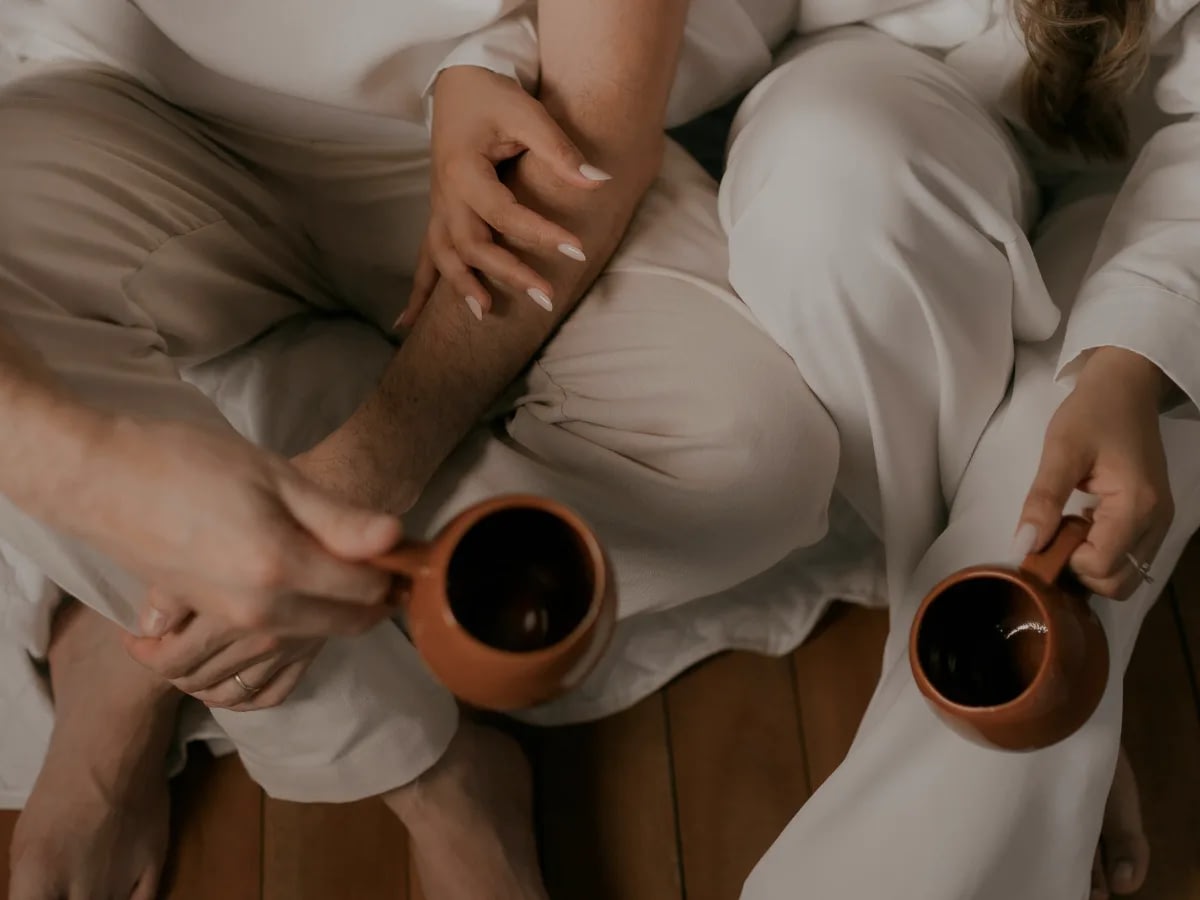 June-25-Blog-ME (1) Couple sitting on a wooden floor in light robes, clasping hands while sharing brown mugs of coffee.