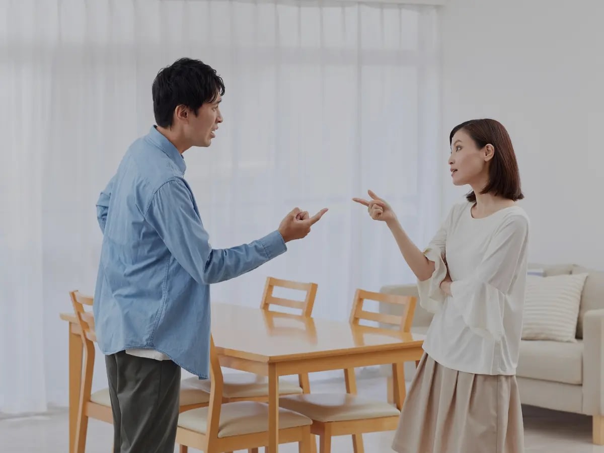 June-25-Blog-ME (1) Couple arguing in a dining room, both are pointing at each other with an argumentative expression.