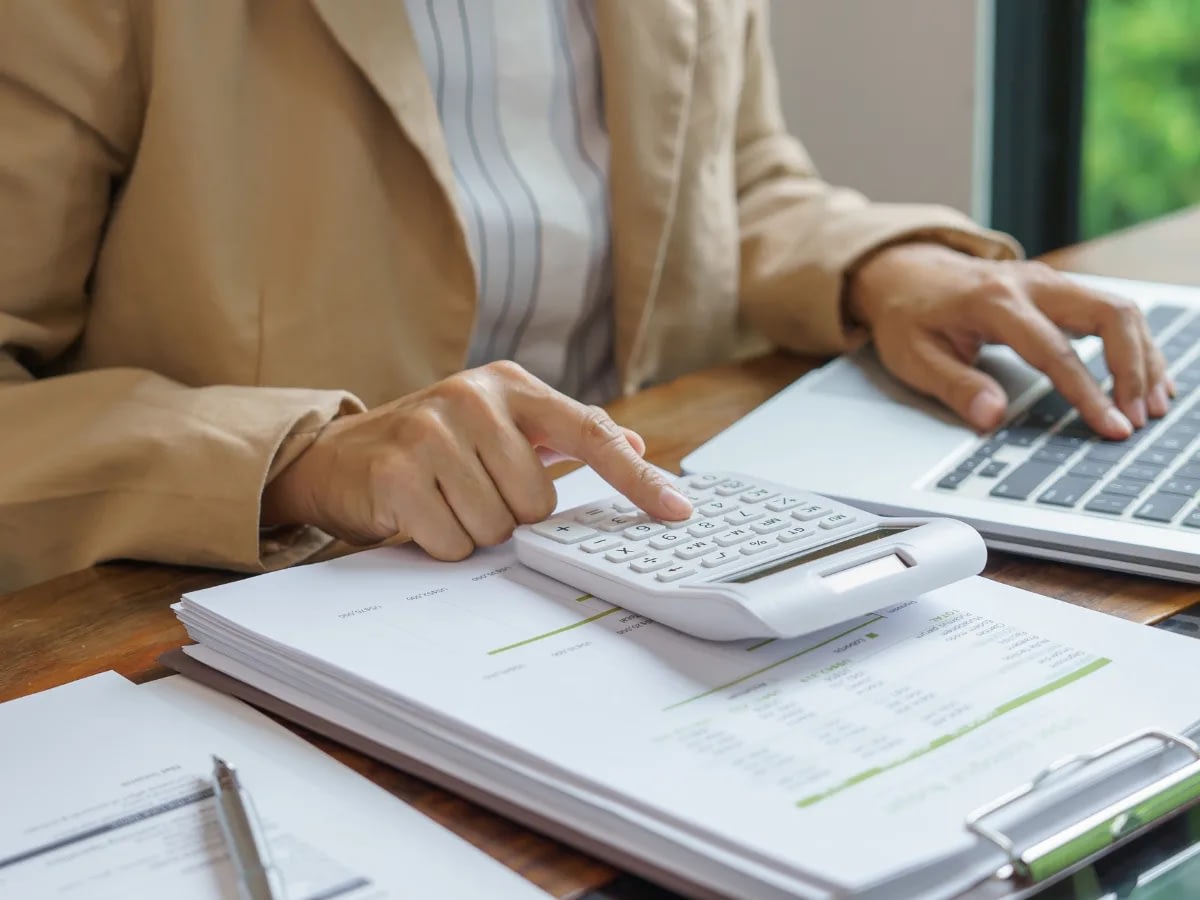Blog July 21 Person in a beige blazer uses a calculator on top of financial documents beside a laptop.