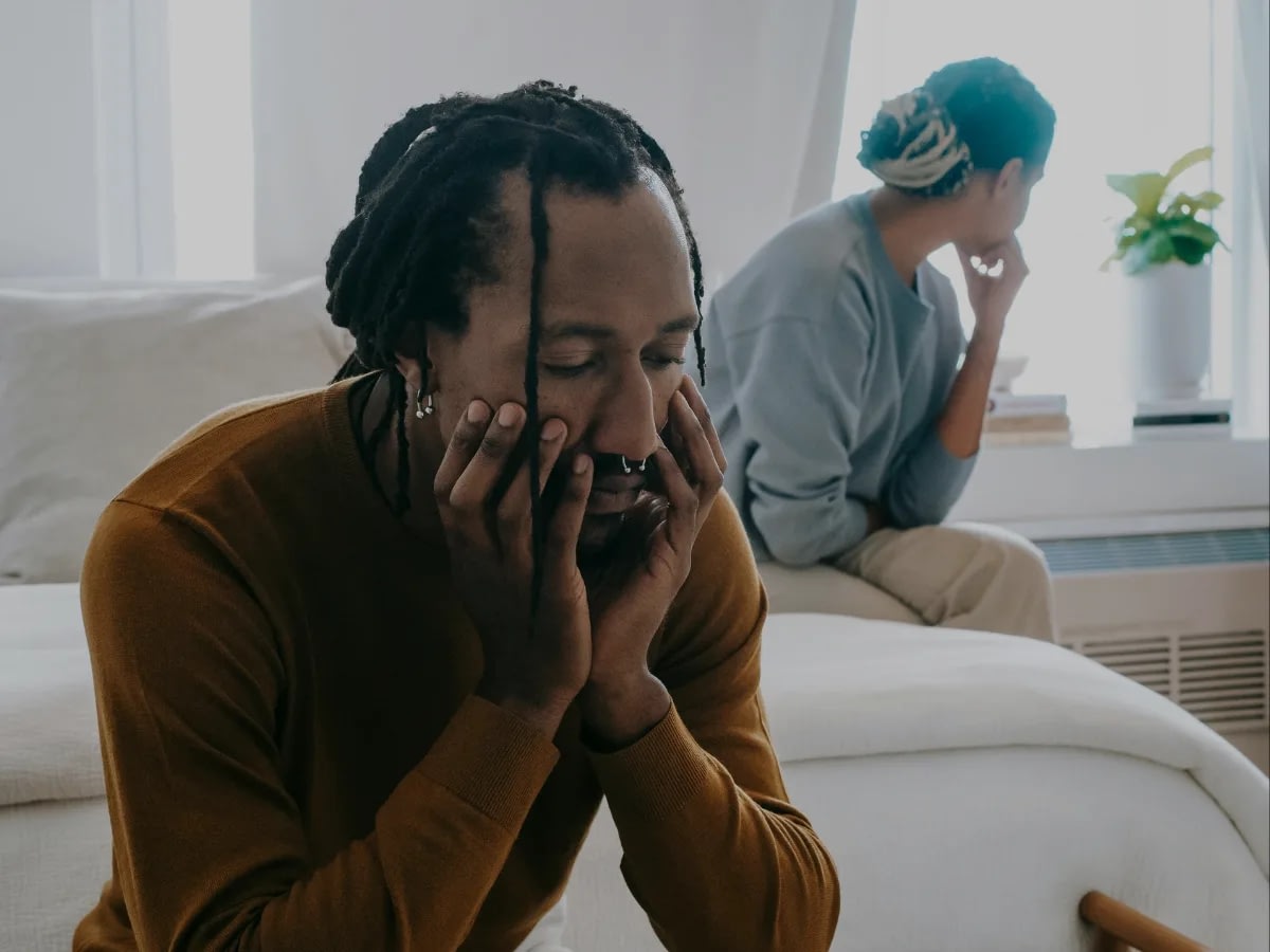 Blog July 23 (2) Man with dreadlocks in a brown sweater sits close to the camera, hands on his face, looking distressed.