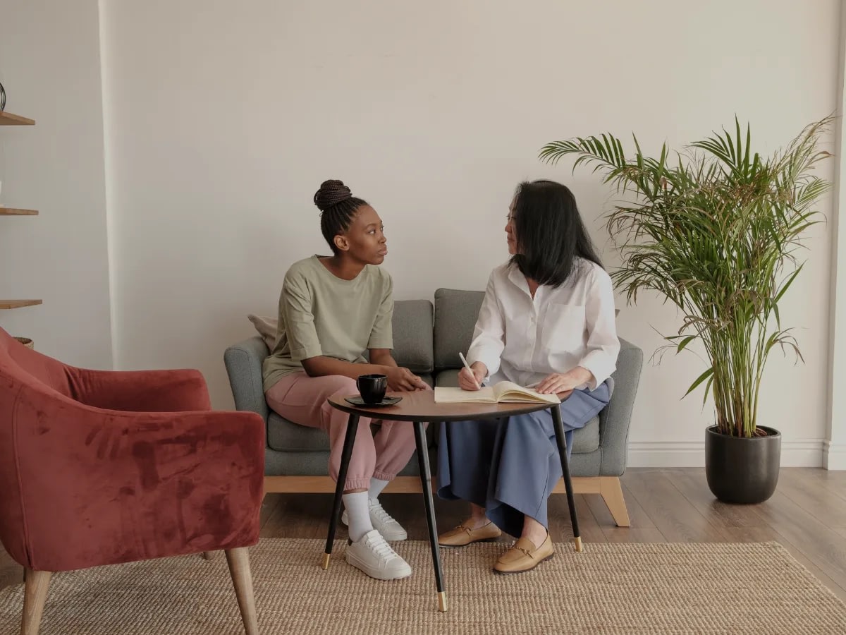 July 14 2 (6) Two women sit on a gray sofa in a living room; one writes in a notebook while the other speaks, with a coffee cup on the table and a tall plant beside them.