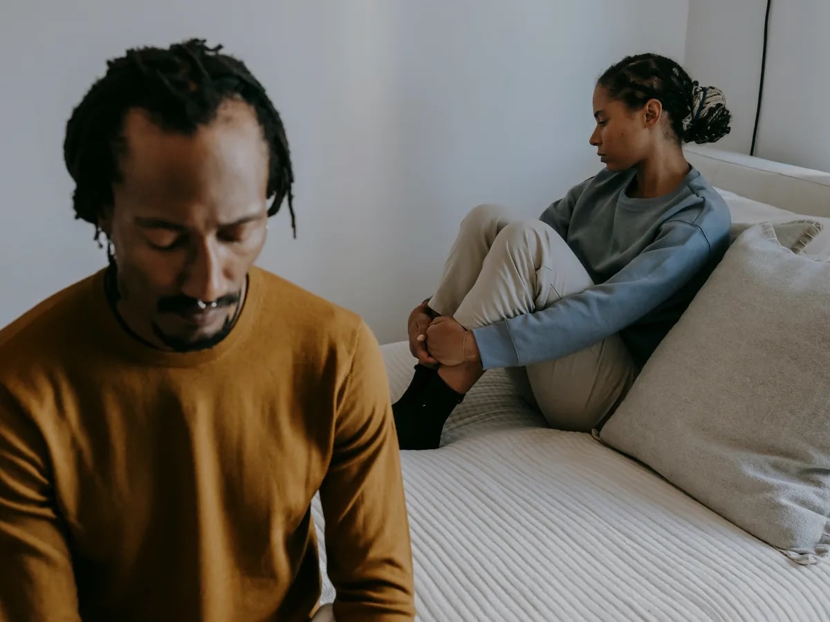 How-to-Make-a-Relationship-Work-With-an-Avoidant-Partner-3 Man in a mustard sweater sits in the foreground with head down; a woman in a blue sweatshirt sits on a bed in the background, knees drawn up and looking away.