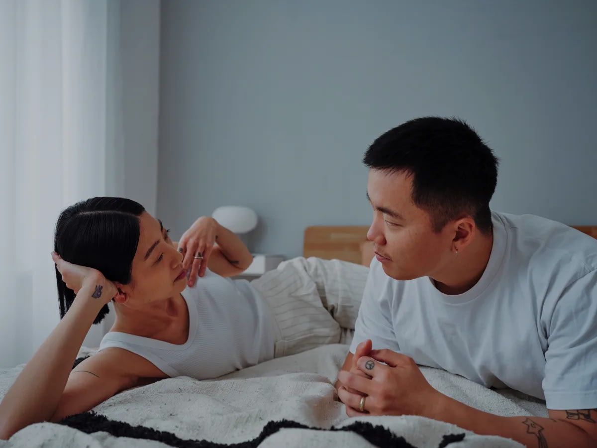 The-Anxious-Avoidant-Trap-Why-One-Partner-Chases-and-the-Other-Withdraws-1 Couple lying on a bed facing each other and talking in a softly lit bedroom, both wearing white tops.