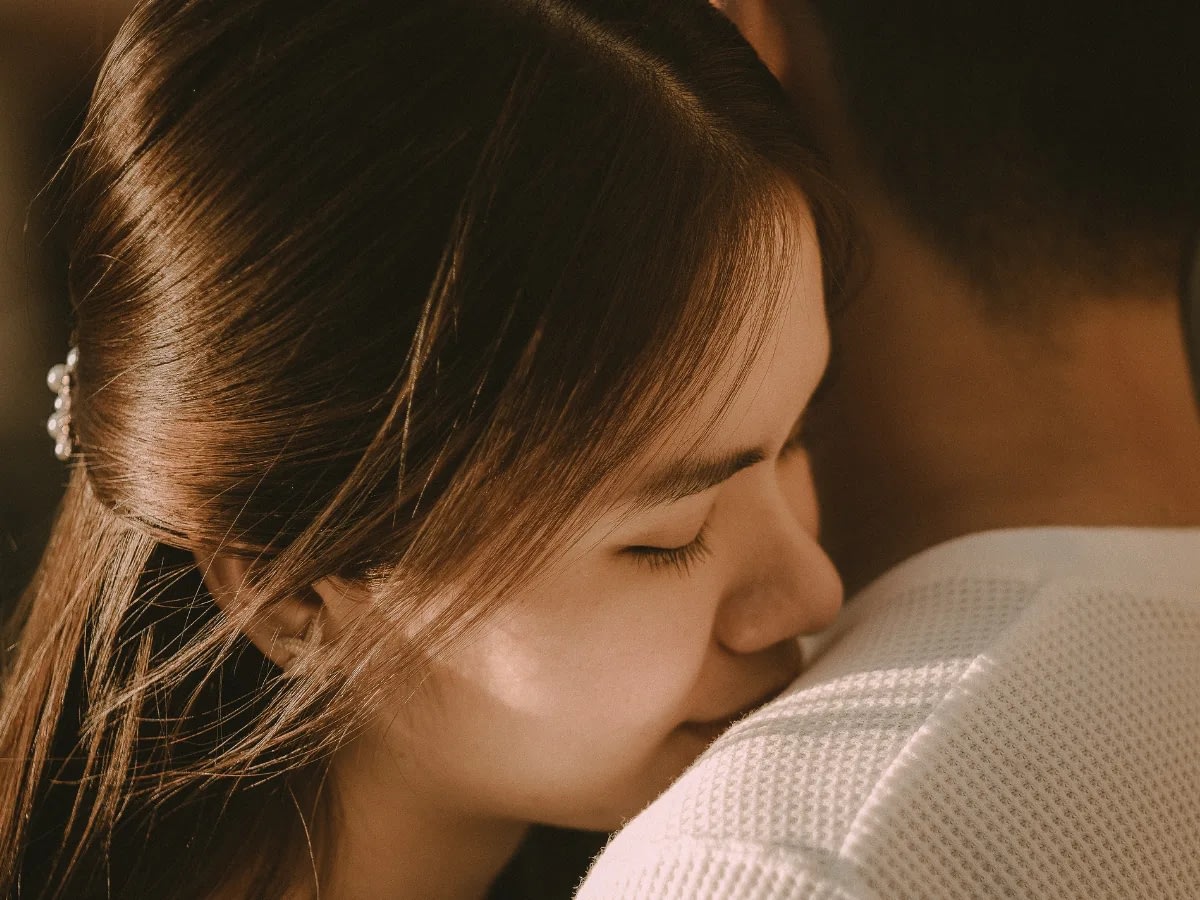 emotional-intimacy-vs-emotional-safety-1 Close-up of a woman with eyes closed resting her head on a man's shoulder, conveying tenderness in warm lighting.