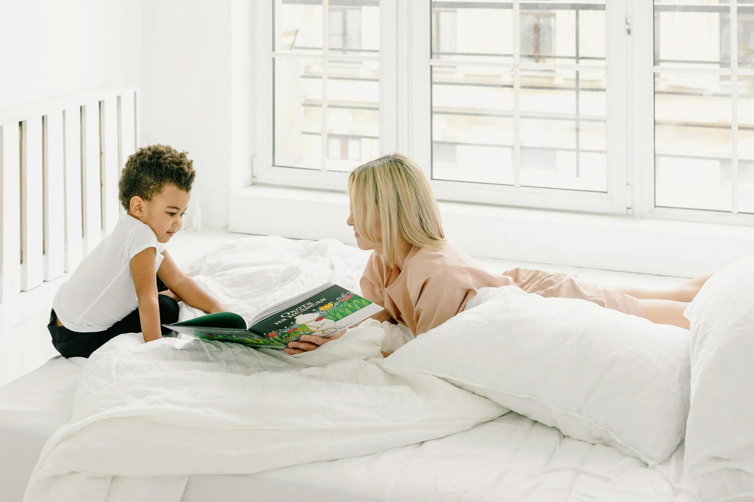 pexels-kseniachernaya-7299837 Woman and child reading a colorful children's book together on a white bed in a bright room with large windows.