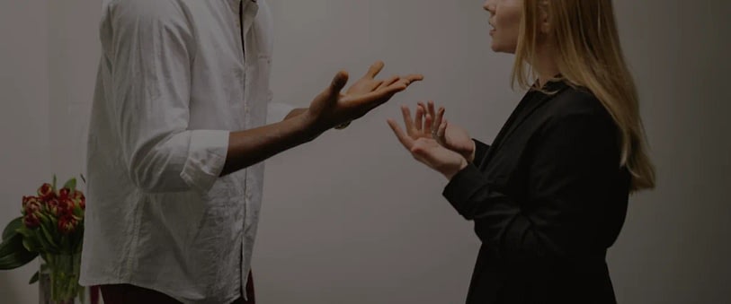 Two professionals converse, using expressive hand gestures, with a vase of red flowers in the background on the left.