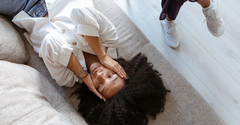 Woman with a big curly afro lying on a beige couch, hands framing her face, a person in white sneakers stands nearby.