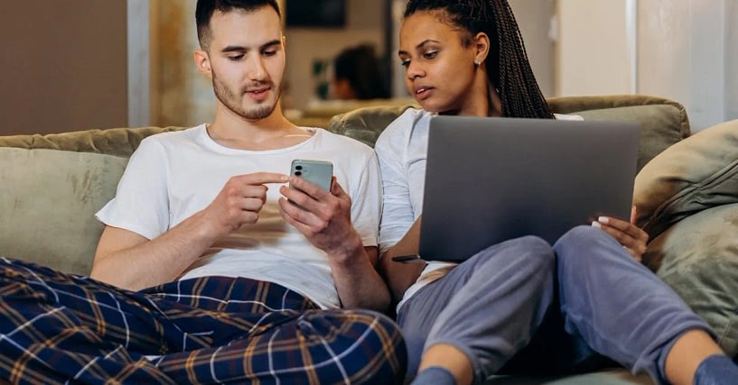 After an affair, a couple is sitting on a couch, the man points at a smartphone while the woman is monitoring the phone while holding a laptop beside him in a living room setting.