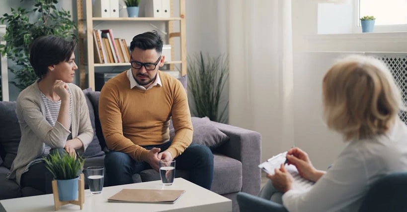 A man and a woman sit on a couch, talking with a clinician who holds a clipboard in a bright living-room therapy session.
