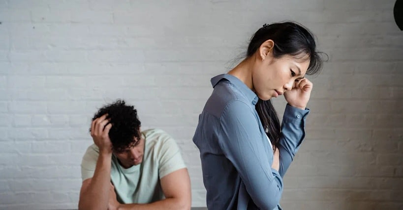 A woman in a blue shirt looks distressed, standing with her hand to her temple while a man sits in the background with his head in his hand, suggesting tension between them.
