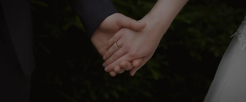 Close-up of a couple holding hands, a physical touch; a wedding ring shows on one finger.